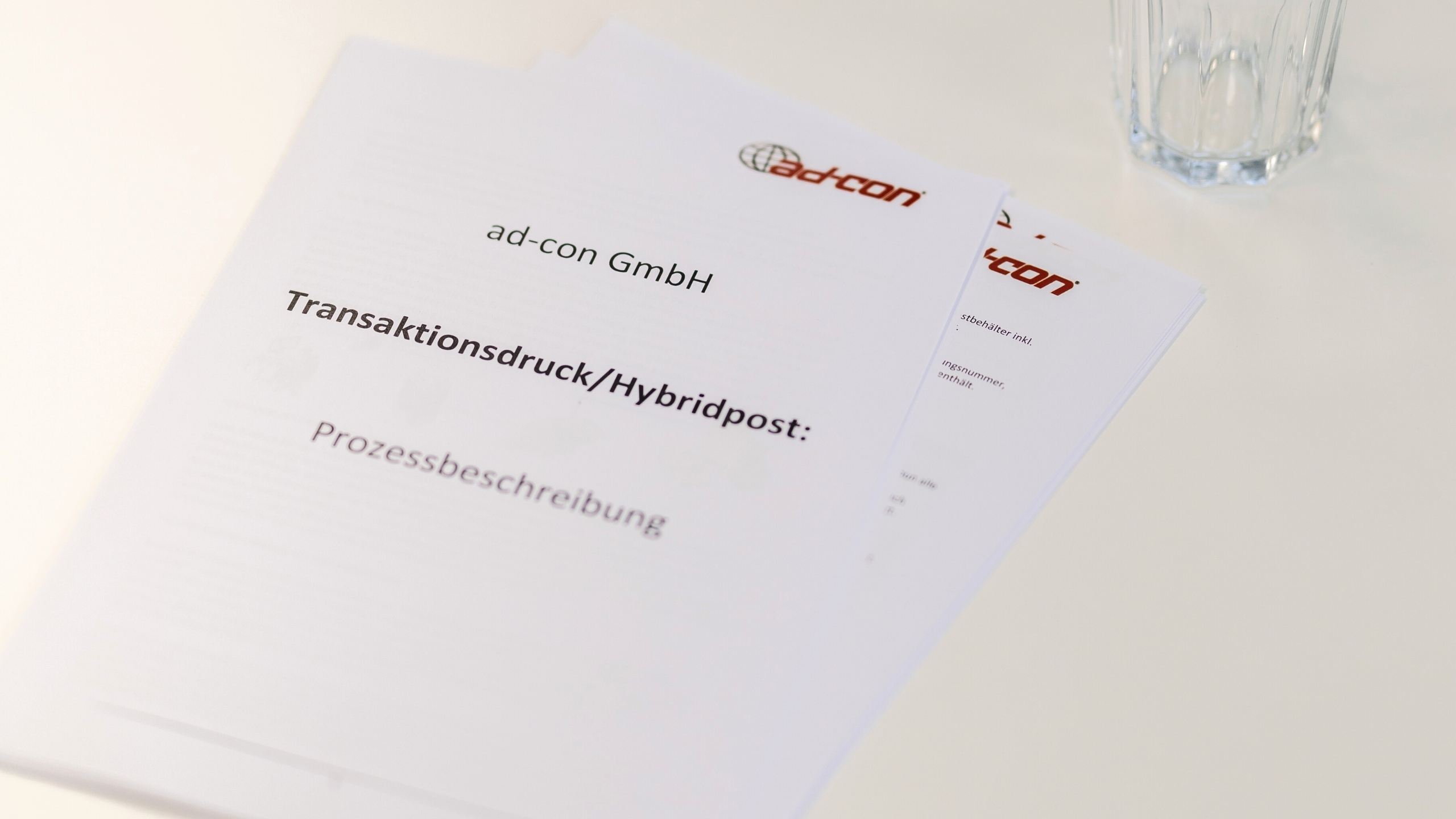 Papierausdrucke liegen auf dem Tisch. Auf der Titelseite ist das ad-con GmbH Logo und der Titel Transaktionsdruck/Hybridpost: Prozessbeschreibung